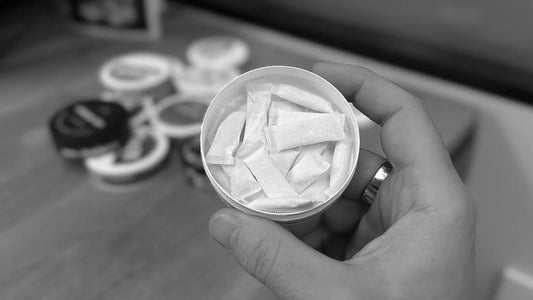 Close-up of an open can of white nicotine pouches held in a hand showing individual pouches with blurred cans on a table
