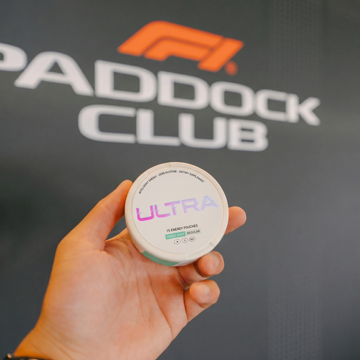 Hand holding Ultra energy pouches tin with holographic ULTRA logo and sweet mint label against blurred Paddock Club backdrop