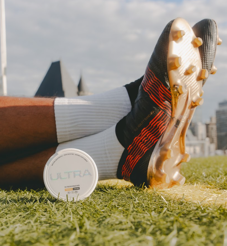 Low angle front view of ULTRA pouch can showing the ULTRA label on turf next to a soccer player’s cleats