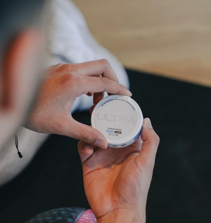 Close-up of hands holding a round can of ULTRA focus pouches showcasing the 15 focus pouches label with a blurred gym floor background